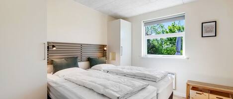 Une chambre moderne avec deux lits, une tête de lit en bois, une table de chevet avec une lampe, une fenêtre donnant sur un paysage verdoyant et un banc en bois avec des tiroirs.