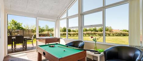A pool table in a room with large windows, a pool table, a pool cue, a pool table chair, and a sofa.