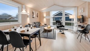Amplia sala de estar con una mesa de comedor, sillas, un sofá y un sillón reclinable. Grandes ventanales ofrecen vistas al exterior.
