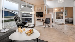 Una sala de estar moderna con sofá, una mesa de comedor con fruta, televisor y una zona de cocina con armarios y televisor.