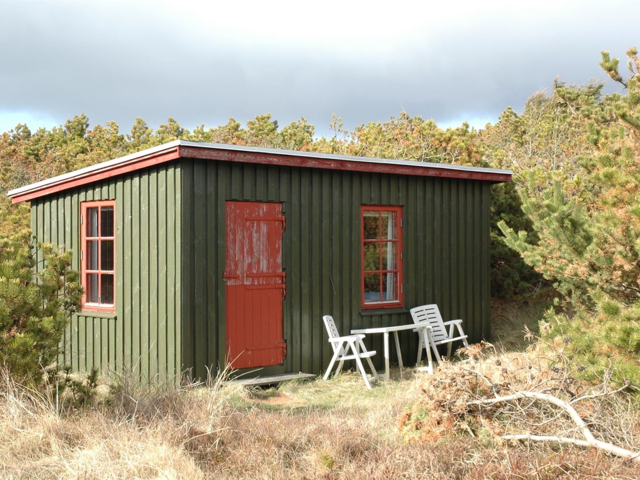 A small, green wooden building with a red door and windows, surrounded by trees and dry grass.