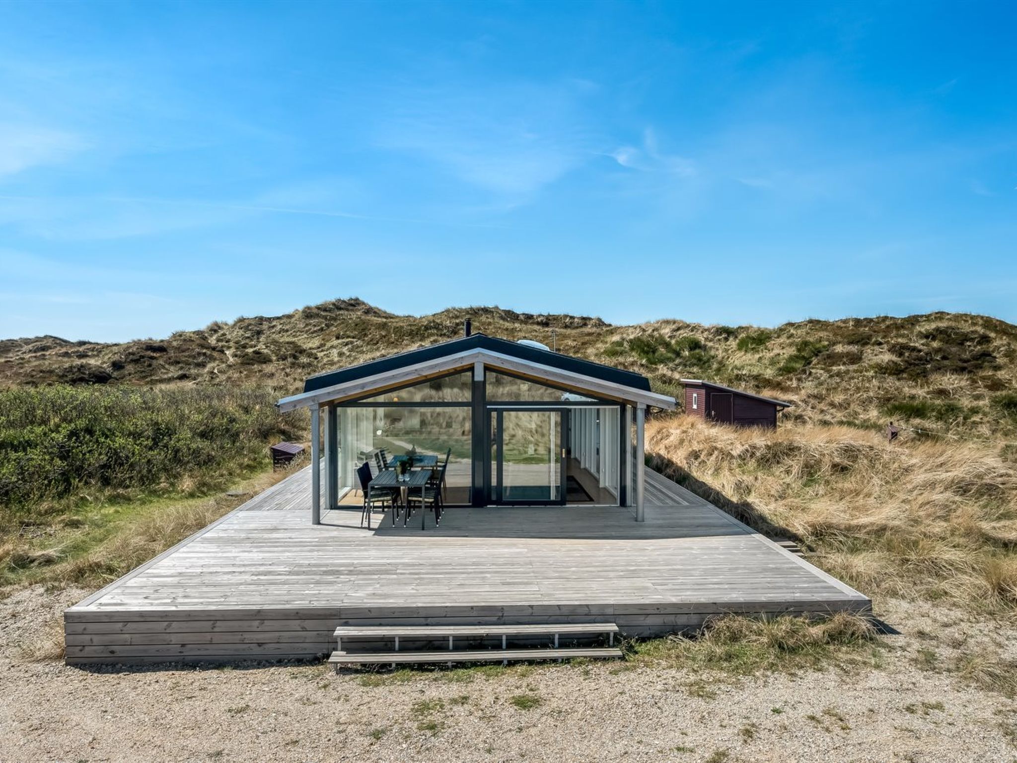 A modern glass-roofed structure with a wooden deck, situated in a natural landscape with dunes in the background.