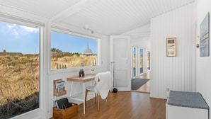 A spacious room with a wooden floor, a desk with a chair, a white bench, and a view of the beach through large windows.