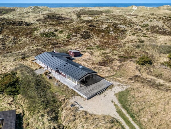 Vista aérea de una casa moderna con techo plano, terraza y una estructura pequeña adyacente, rodeada de dunas y cielo despejado.