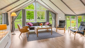 Une pièce spacieuse avec un plafond haut, de grandes fenêtres, un canapé, des fauteuils, une table basse et une lampe.