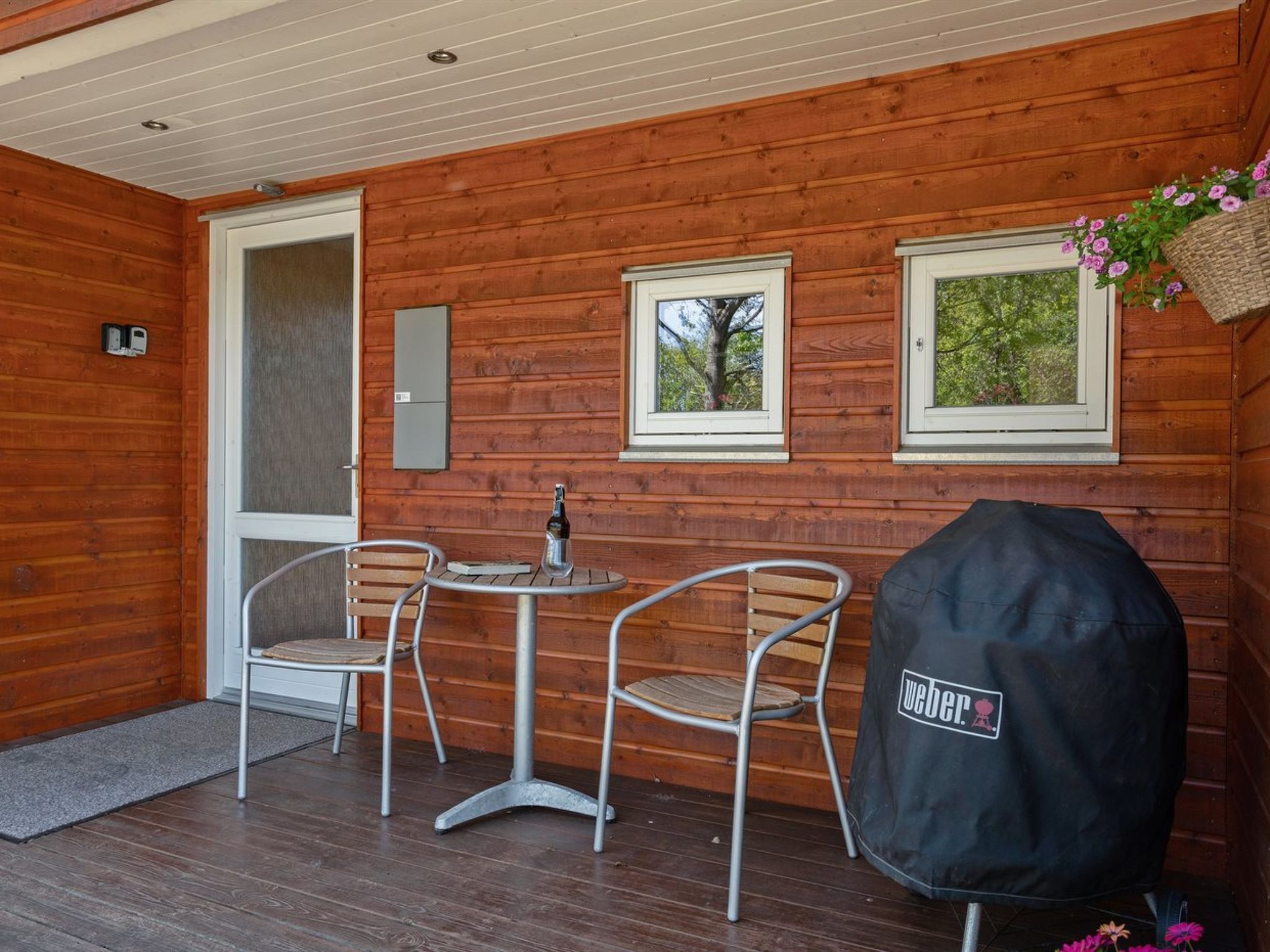 A wooden cabin with a small patio area featuring two metal chairs, a table, and a grill cover.