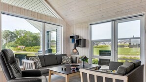 Una sala de estar moderna con sofá, sillón y mesa, con ventanas amplias que ofrecen vistas al exterior.