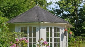 A small, white gazebo with a dark roof, surrounded by lush greenery and pink flowers.