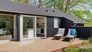 A modern house with a wooden deck, large sliding glass doors leading to a patio, and a small outdoor seating area with two chairs.