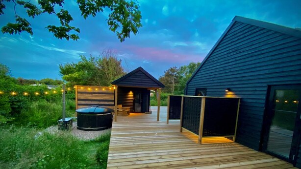 A wooden deck leading to a modern building with a hot tub, a covered seating area, and string lights.