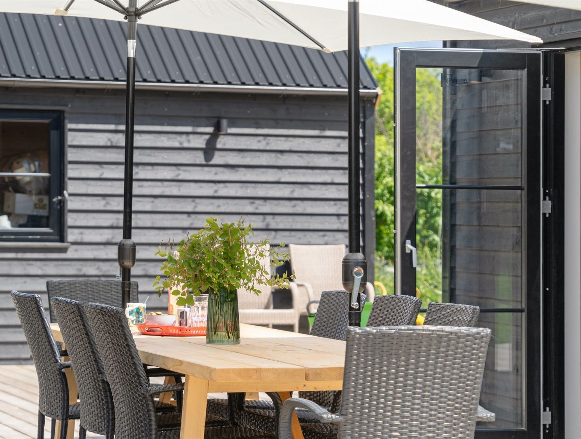 An outdoor dining area with a wooden table and wicker chairs, a potted plant, and a grill.