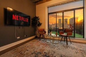 A room with a large TV displaying 'NETFLIX,' a plant, two chairs, a small table, and a view of a sunset through a large window.