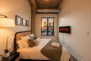 A hotel room with a bed, a bedside lamp, a wall-mounted TV, a window with a city view, and a potted plant.