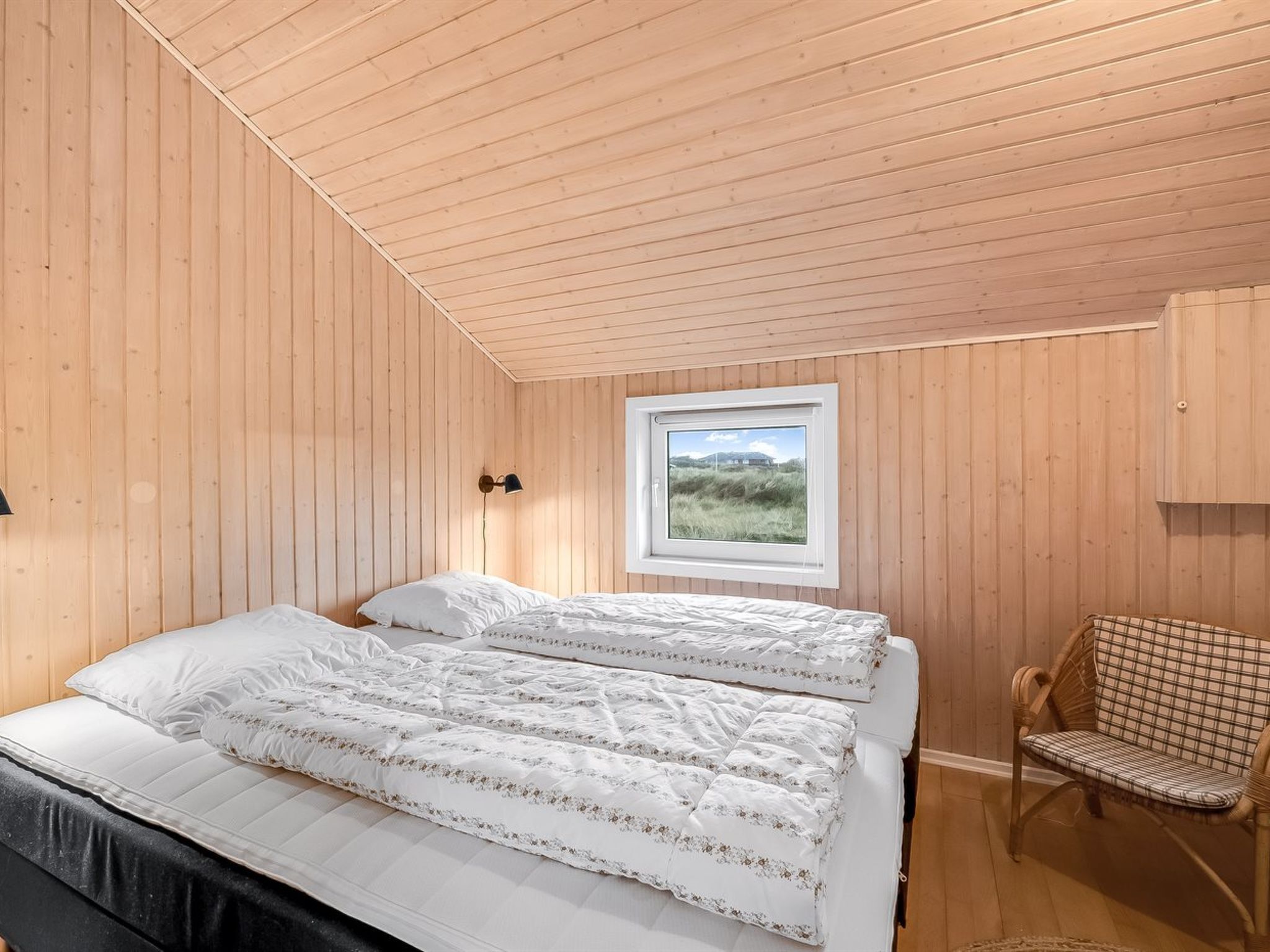 Une chambre avec un plafond en bois, une fenêtre donnant sur l’extérieur, un lit avec une literie blanche et un fauteuil en osier.