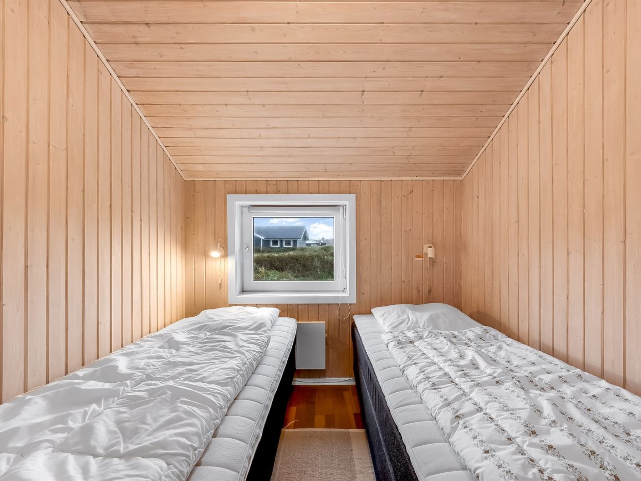 A small, wooden-paneled room with two beds, a window, and a view of a house outside.