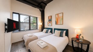 Una camera d'albergo con due letti, una TV a parete, una finestra e un tavolino con una lampada.