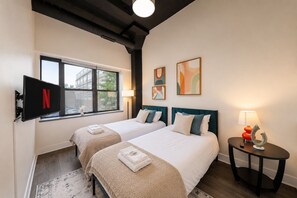 A hotel room with two beds, a TV mounted on the wall, a window, and a small table with a lamp.