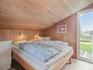 A bedroom with a wooden ceiling, a bed with white bedding, a nightstand, a wall-mounted lamp, and a window with a view of greenery.