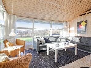 Un salon moderne avec un canapé gris, une table basse blanche, des fauteuils en osier et de grandes fenêtres donnant sur l’extérieur.