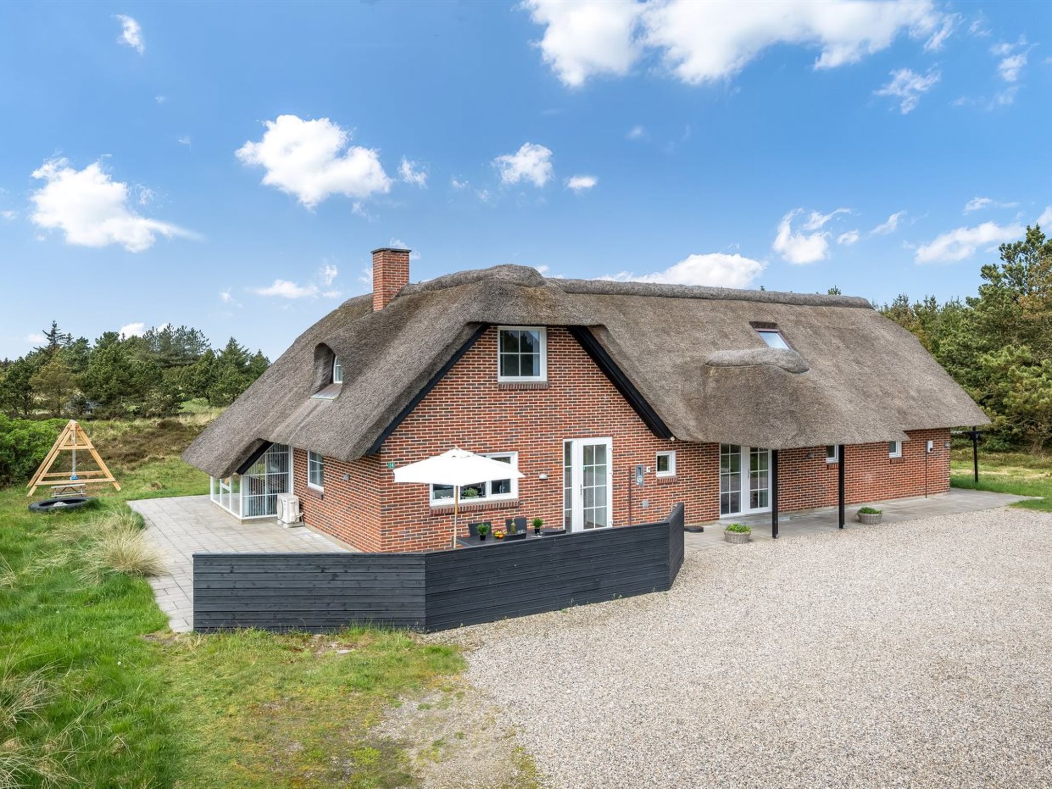 A thatched-roof house with a brick exterior, a gravel driveway, and a wooden swing set in the yard.