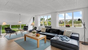A spacious living room with a black leather sofa, a wooden coffee table, and a dining area with chairs and a table. Large windows offer views of the outdoors. - "Vangel" - 500m from the sea by Interhome (Blåvand)