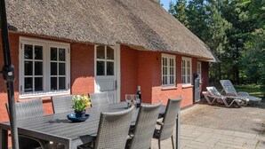 A thatched-roof house with a dining table and chairs, a small garden, and a pool area.