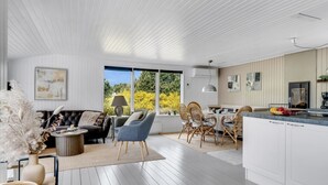 A modern kitchen with white cabinetry and a large island. There is a dining area with wicker chairs and a sofa. The room has large windows with a view of greenery outside. - "Noralee" - 3.5km from the sea by Interhome (Blokhus)