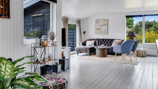 A spacious living area with a dark-colored sofa, a blue armchair, and a round ottoman. There is a wooden floor, a large window, and a ceiling with white panels. - "Noralee" - 3.5km from the sea by Interhome (Blokhus)