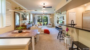 A spacious living area with a sofa, coffee table, and a dining table. There is a kitchen area with a refrigerator, sink, and bar stools. The room has a ceiling fan and large windows. - Dream of Aloha (Kapolei)