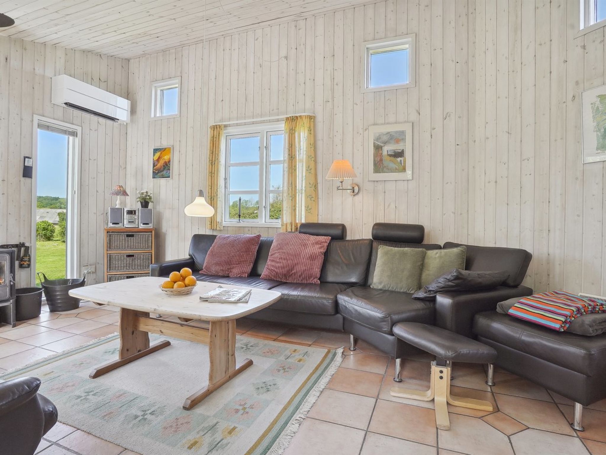 A cozy living room with a wooden ceiling, a black leather sofa, a wooden coffee table, and a view of the outdoors through large windows.