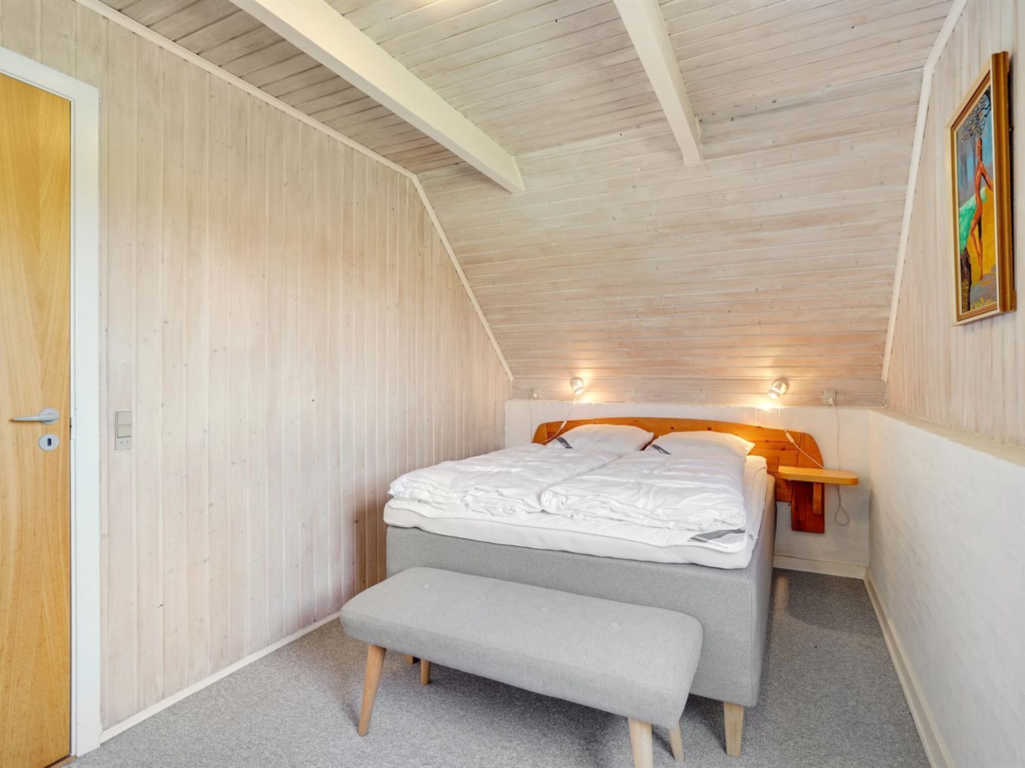 Une chambre avec un plafond en pente, des murs en bois, un lit avec une literie blanche, un banc et une table de chevet avec une lampe.