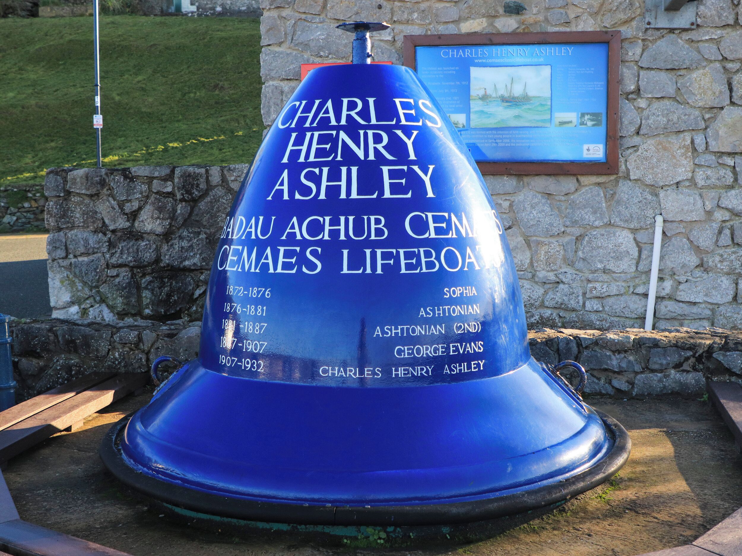 A blue conical monument with inscriptions, including names and dates, set against a stone wall with a plaque in the background.