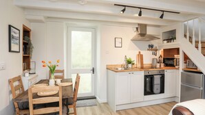 A modern kitchen with white cabinets, a wooden dining table, and a built-in oven. - Bwthyn Lili (Cemaes Bay)