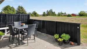 Eine Außenpatio mit Tisch und Stühlen, umgeben von einem schwarzen Zaun, mit Topfpflanzen und Blick auf ein Feld sowie Gebäude im Hintergrund.