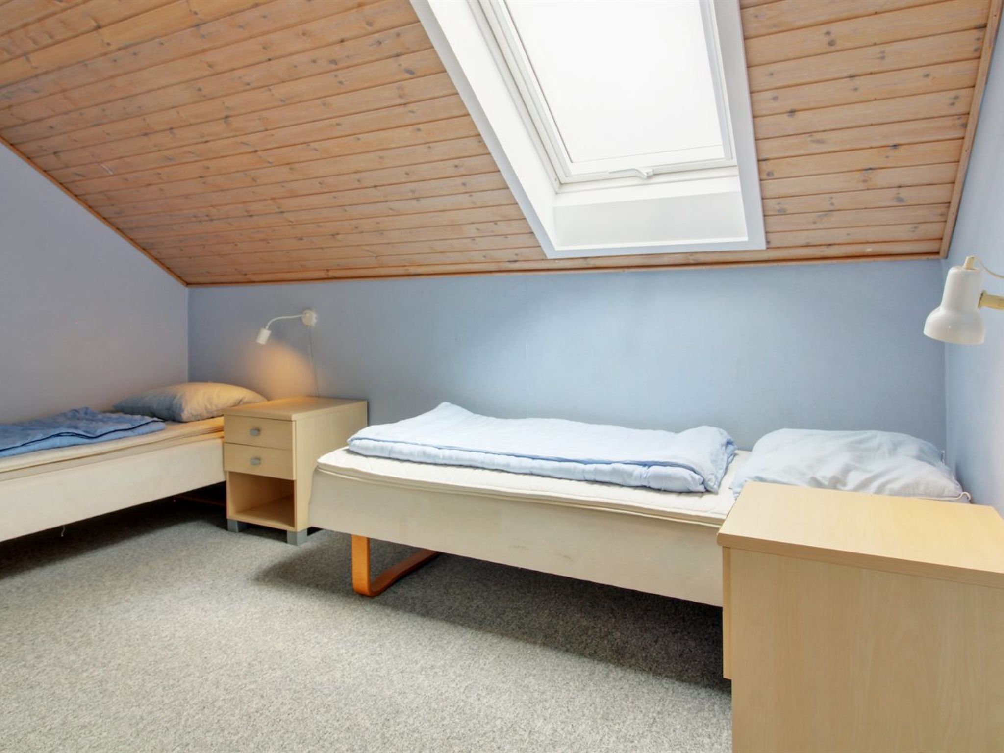 Quarto em dormitório com duas camas, um claraboia e uma escrivaninha com luminária.