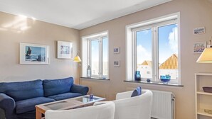A modern living room with a blue sofa, white armchairs, a glass coffee table, and large windows offering a view of buildings and a clear sky. - "Gerrit" - 450m from the sea by Interhome (Løkken)