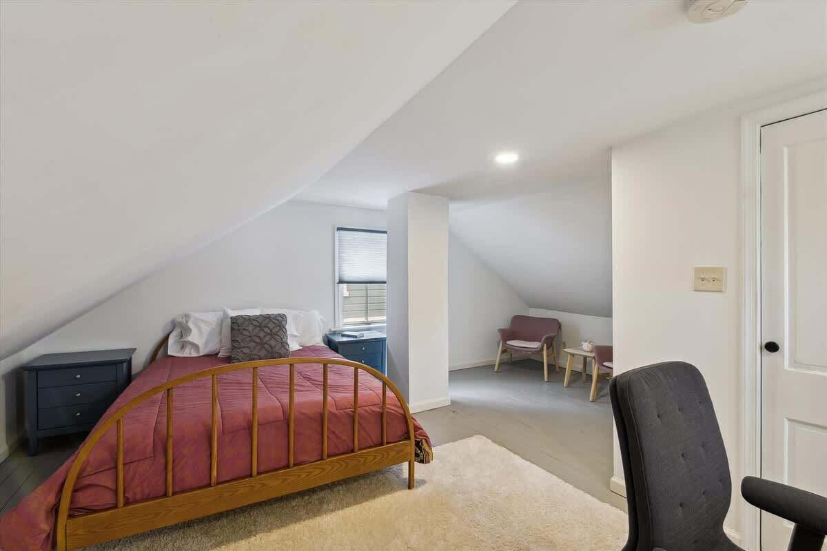 A bedroom with a sloped ceiling, a bed with a wooden frame, a bedside table, a chair, and a small table with a chair.
