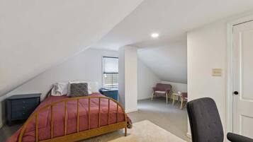A bedroom with a sloped ceiling, a bed with a wooden frame, a bedside table, a chair, and a small table with a chair.