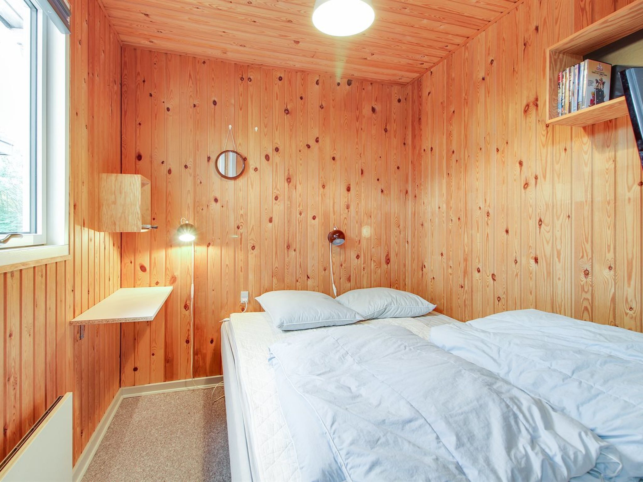 A wooden-paneled room with a bed, a bedside table, a lamp, and a shelf with books.