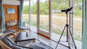 Een kamer met houten plafond, grote schuifdeuren, een telescoop op statief, een tafel met stoelen en een ledikant met babystoel.