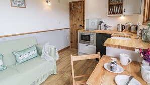 A cozy room with a wooden floor, a green sofa with decorative pillows, a wooden dining table with plates and a cup, and a kitchenette with a stove, oven, and sink. - Tan Y Castell Shepherds Hut (Llangollen)