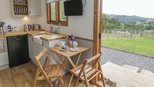 A compact kitchen with a wooden table and chairs, a built-in dishwasher, and a large window offering a view of a grassy landscape. - Tan Y Castell Shepherds Hut (Llangollen)