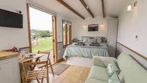 A cozy room with a bed, a sofa, a wooden table, and a chair. There is a window with a view of greenery outside. - Tan Y Castell Shepherds Hut (Llangollen)