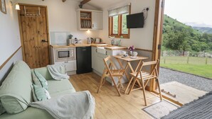 A cozy room with a wooden floor, a green sofa, a dining table with chairs, a kitchen area with a microwave, a coffee maker, and a TV mounted on the wall. - Tan Y Castell Shepherds Hut (Llangollen)