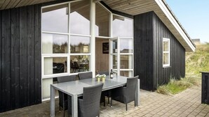 Moderni ulkotila, jossa on ruokapöytä ja -tuolit, suuret ikkunat ja puinen julkisivu.