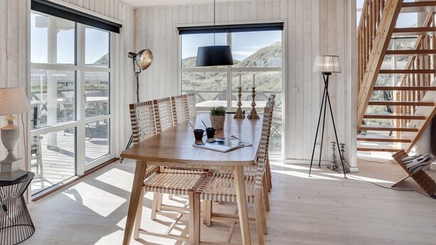 Een ruime eethoek met een houten tafel en stoelen, grote ramen met uitzicht op de omgeving, en een trap met houten leuningen.