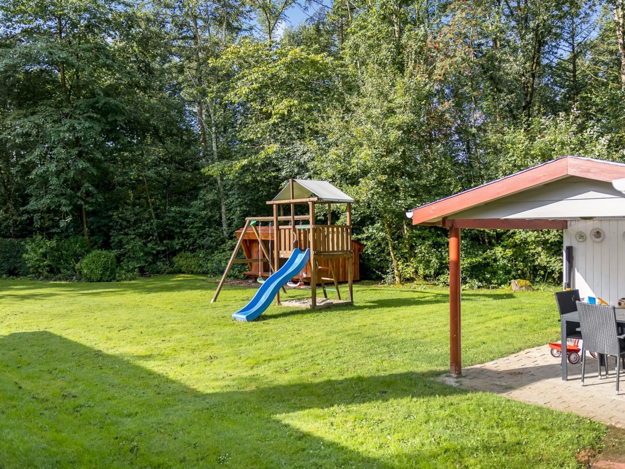 A wooden play structure with a slide in a grassy backyard surrounded by trees.