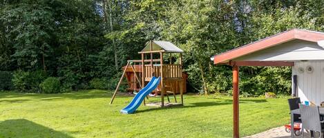 A wooden play structure with a slide in a grassy backyard surrounded by trees.