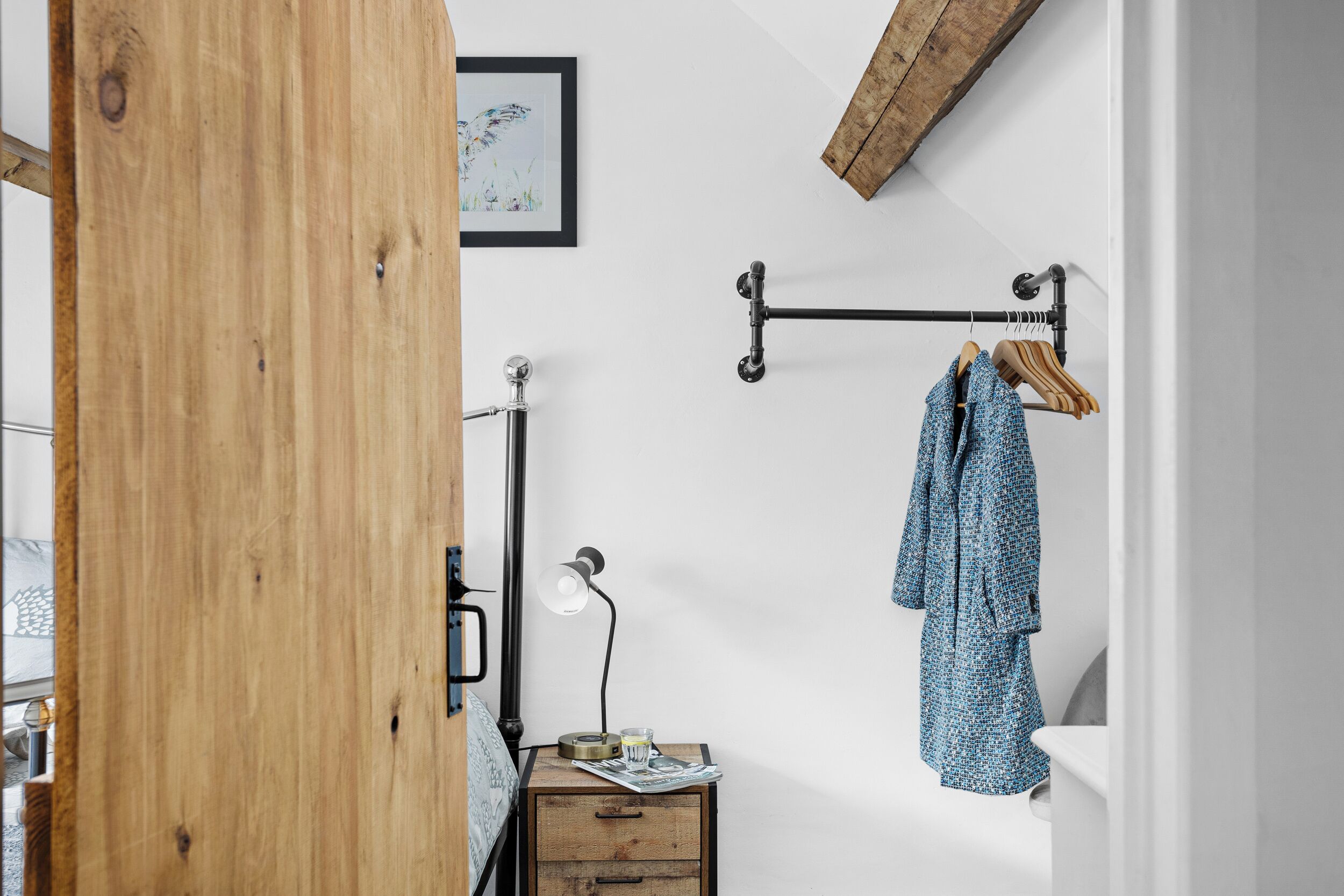 A bedroom with a wooden door, a bedside table with a lamp, a framed picture on the wall, and a coat rack with a coat.