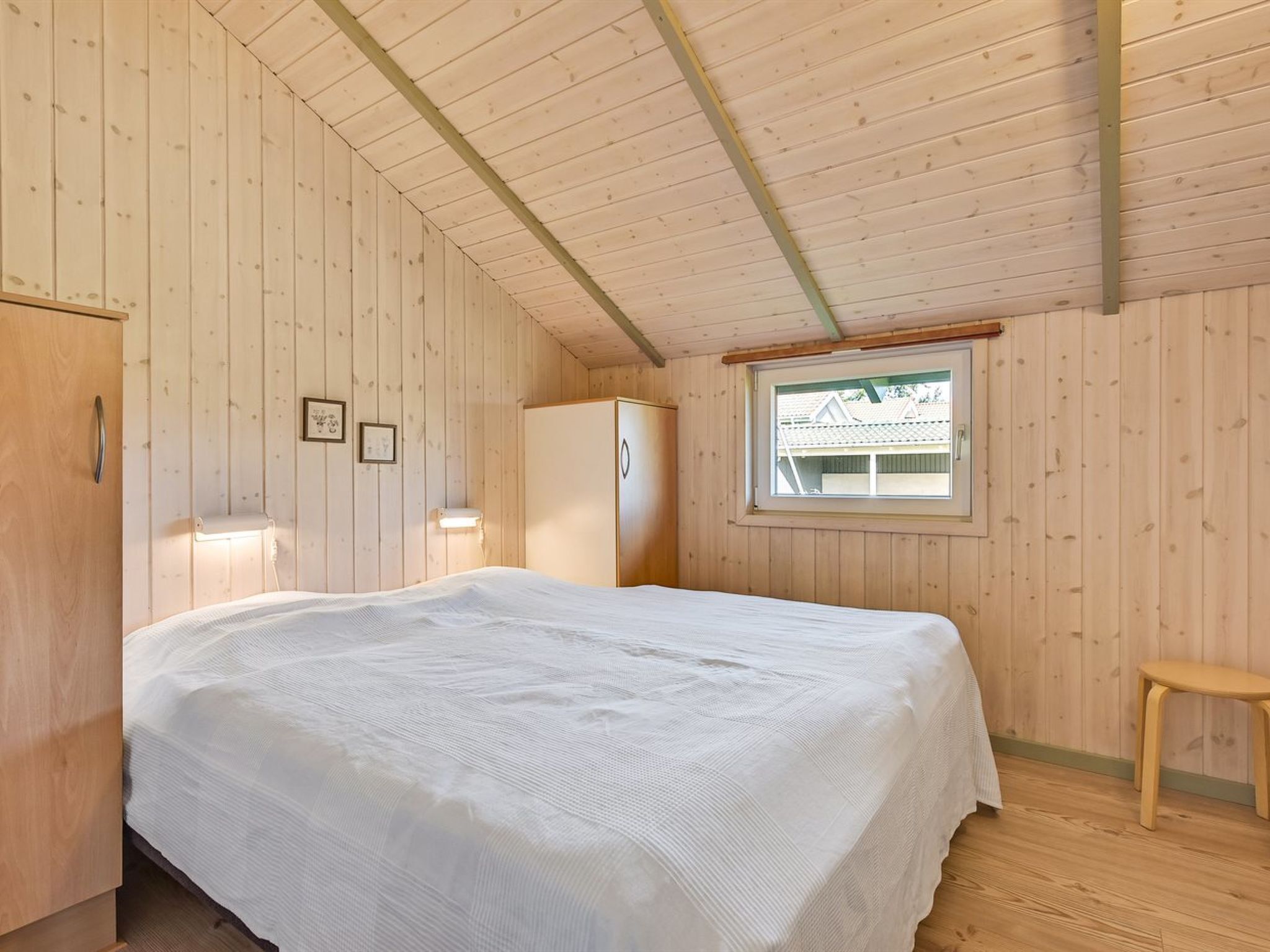 Een kamer met houten lambrisering, een groot bed, een raam en een houten kruk.